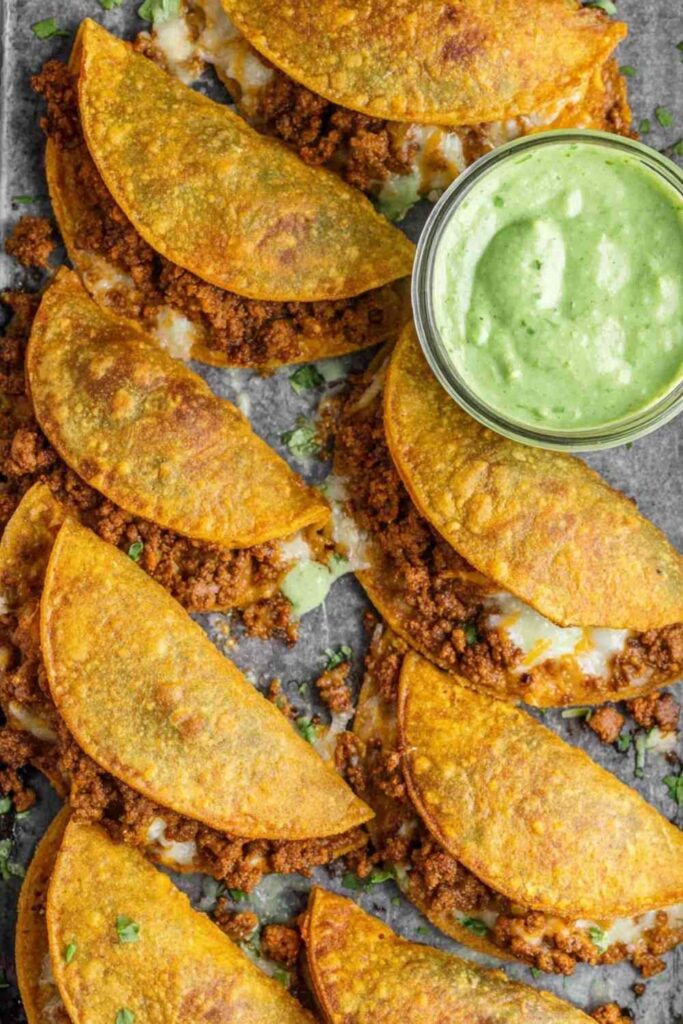 Crispy chicken tacos baked golden on a white plate served with avocado cilantro sauce and lime wedges