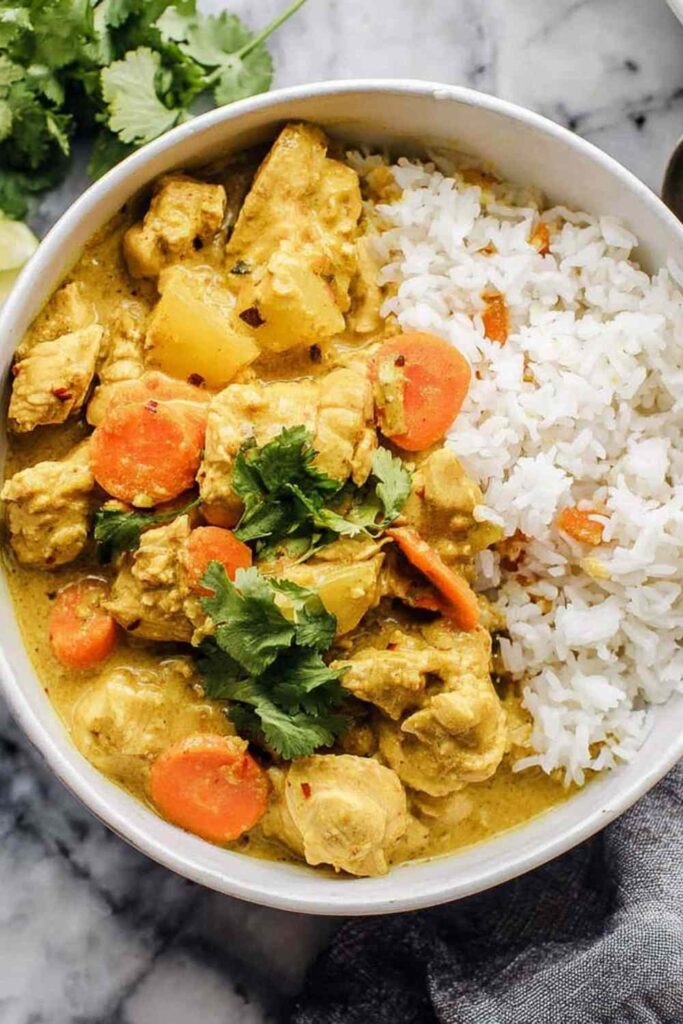 Crockpot curry chicken served over basmati rice in a white bowl topped with fresh cilantro and chopped cashews