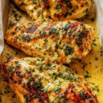 Garlic butter baked chicken breast served on a white plate with fresh parsley and lemon wedges