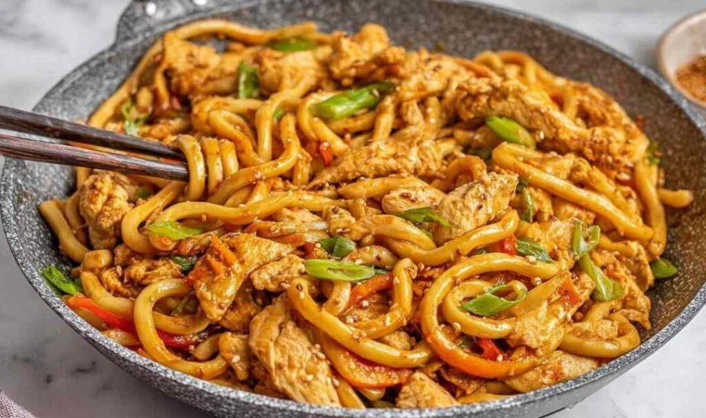 Ginger chicken udon noodles stir fry in a skillet topped with sesame seeds and scallions