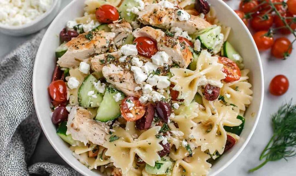 Greek chicken pasta salad with feta cheese, Kalamata olives, cucumber, and cherry tomatoes in a large white bowl