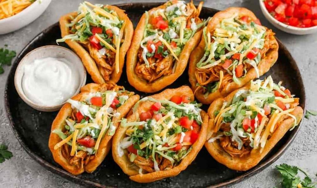 Crispy homemade chicken chalupas filled with seasoned chicken, shredded cheese, lettuce, and diced tomatoes on a wooden serving board