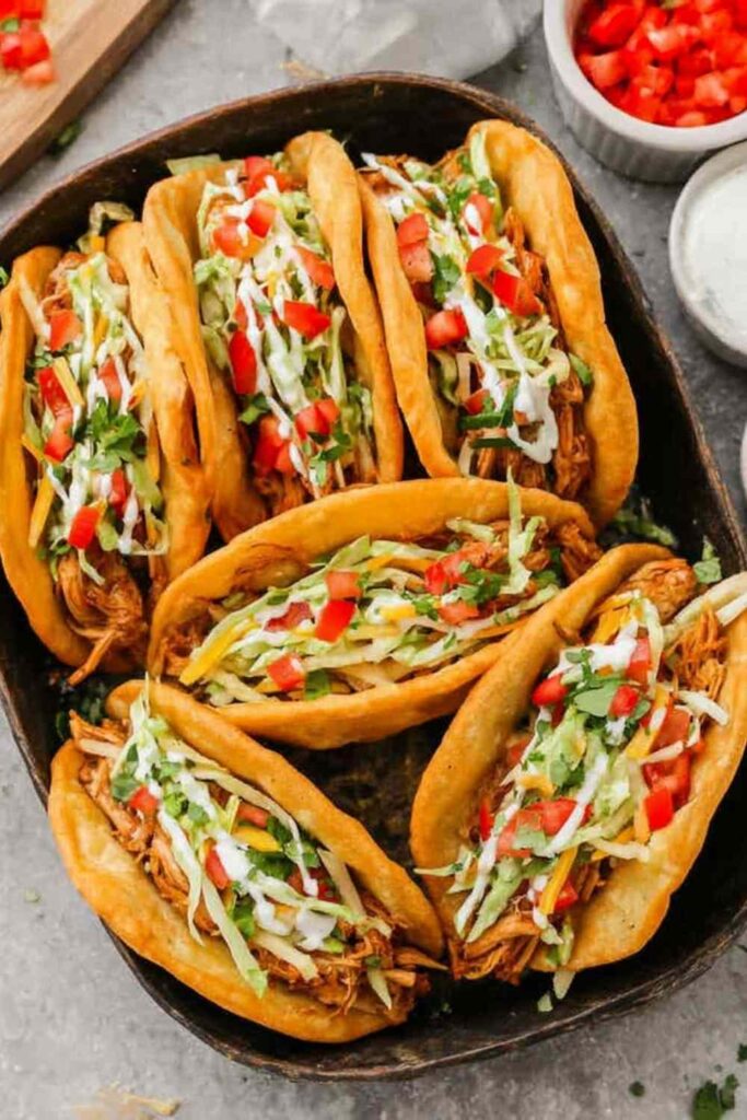 Crispy homemade chicken chalupas filled with seasoned chicken, shredded cheese, lettuce, and diced tomatoes on a wooden serving board