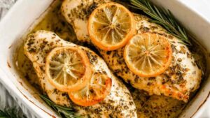Golden seared lemon rosemary chicken breasts in a white baking dish with fresh lemon slices and rosemary sprigs