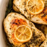 Golden seared lemon rosemary chicken breasts in a white baking dish with fresh lemon slices and rosemary sprigs