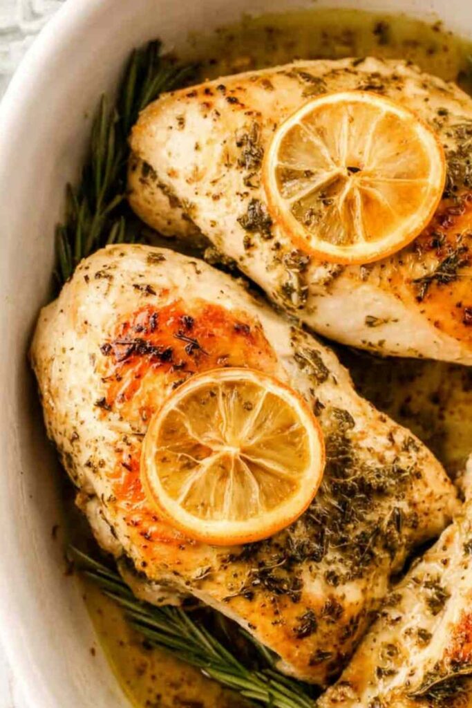 Golden seared lemon rosemary chicken breasts in a white baking dish with fresh lemon slices and rosemary sprigs
