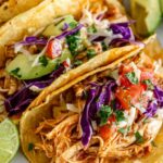 Rotisserie chicken tacos topped with shredded red cabbage, avocado slices, cheddar cheese, and fresh cilantro on warm corn tortillas