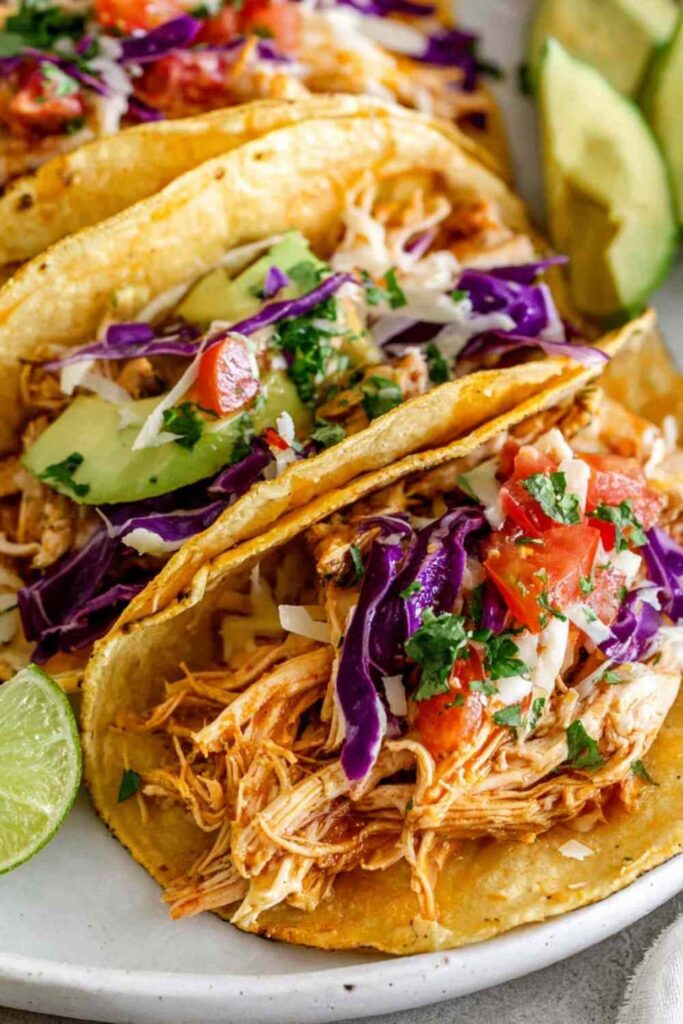 Rotisserie chicken tacos topped with shredded red cabbage, avocado slices, cheddar cheese, and fresh cilantro on warm corn tortillas