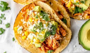 Shredded chipotle chicken tacos on charred corn tortillas topped with avocado, sweet corn, cilantro, and cotija cheese