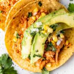 Shredded chipotle chicken tacos on charred corn tortillas topped with avocado, sweet corn, cilantro, and cotija cheese