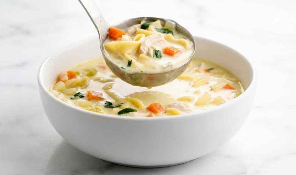 A bowl of creamy chicken noodle soup with shredded chicken, egg noodles, diced carrots, and fresh parsley garnish