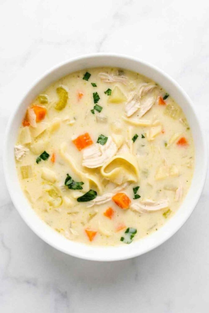 A bowl of creamy chicken noodle soup with shredded chicken, egg noodles, diced carrots, and fresh parsley garnish