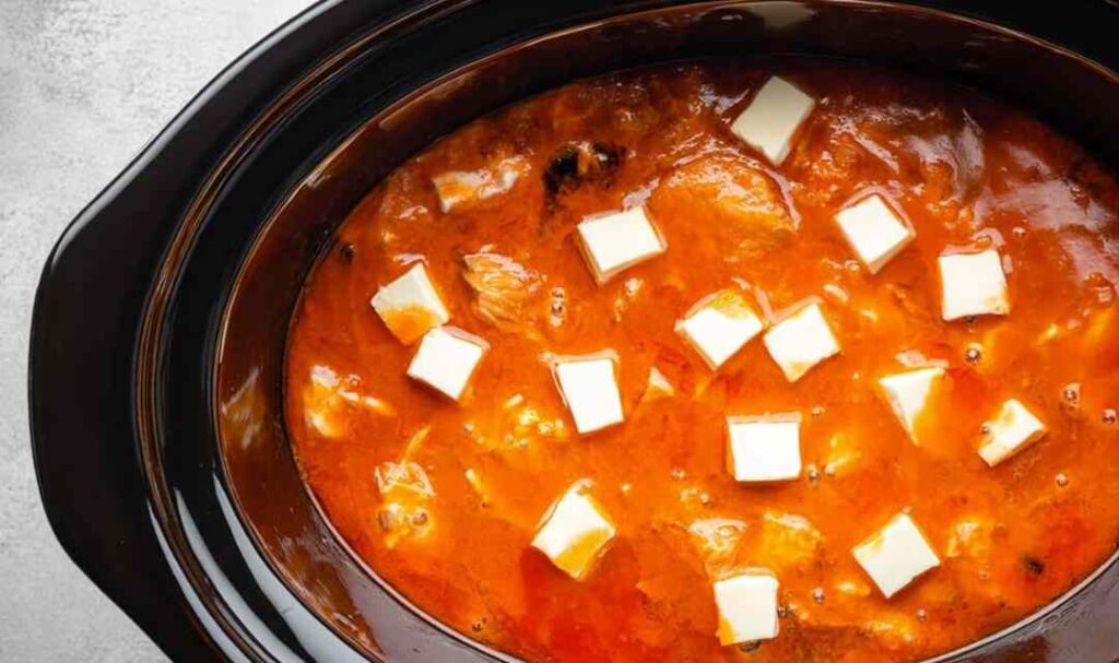 Buffalo sauce being poured over ranch seasoned chicken in a crockpot with cubed butter on top