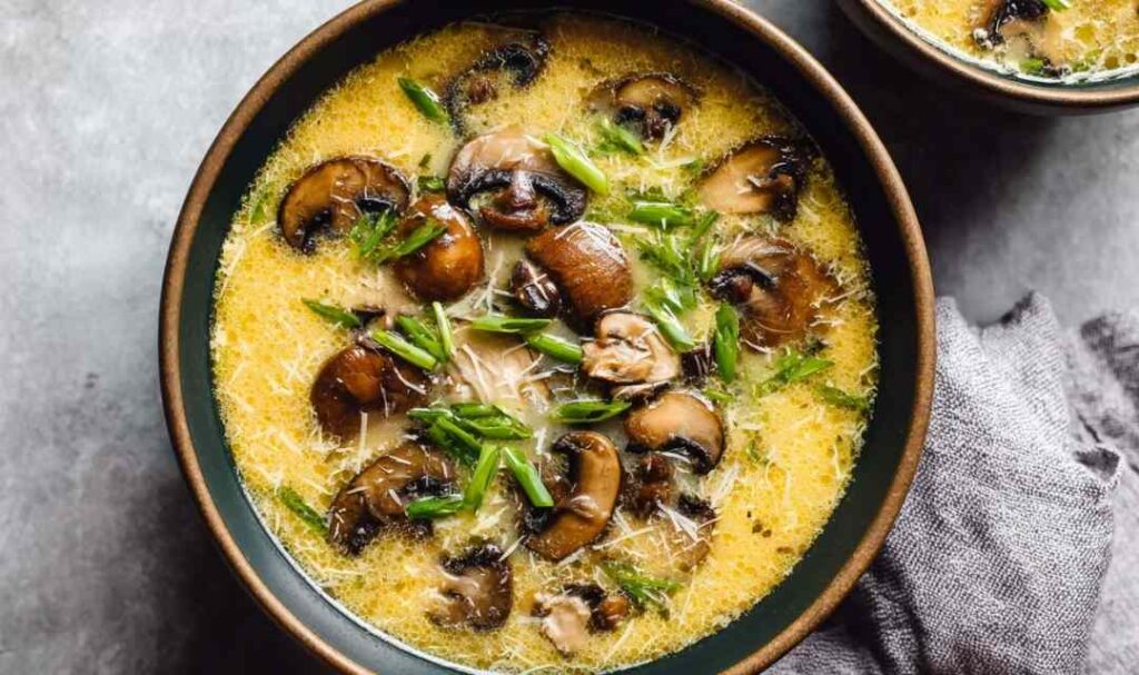 A bowl of homemade egg drop soup with silky egg ribbons, sliced cremini mushrooms, and chopped green onions in a golden chicken broth