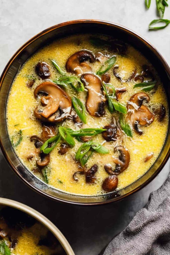 A bowl of homemade egg drop soup with silky egg ribbons, sliced cremini mushrooms, and chopped green onions in a golden chicken broth